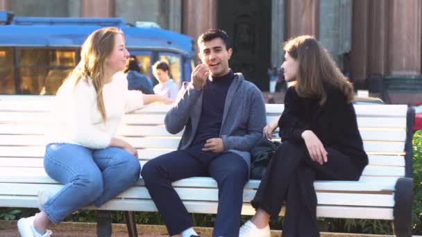 Két fiatal lány és egy férfi barát ül egy padon a nyári parkban, és boldogan beszélgetnek. A média. Két mosolygó nő és egy férfi portréja, akik együtt töltik az időt és beszélgetnek.. — Stock videók