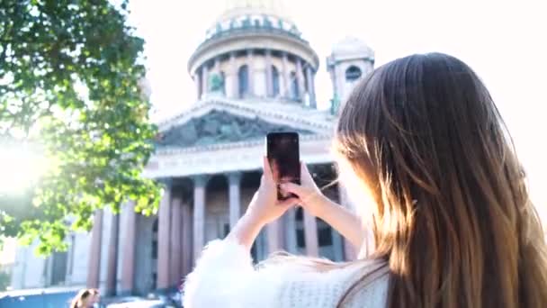 Jonge vrouw op vakantie fotograferen met haar smartphone een prachtig historisch gebouw. Concept. Achteraanzicht van een jonge toerist die staat en een smartphone gebruikt om foto 's te maken van de Saint Isaac — Stockvideo