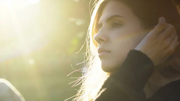 Woman face close up view at golden sunset. Media. Side view of young beautiful woman in a knitted black sweater in a sunny evening tucking her hair behind ear.