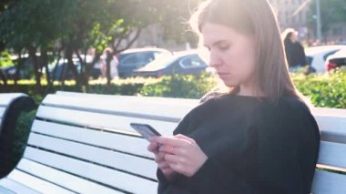 Akıllı telefon kullanarak yaz güneşinin parlak ışınları altında genç bir bayanın portresi. Medya. Şehir parkında beyaz bir bankta oturmuş mesaj atan güzel bir kadın..