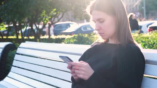 Portret van een jonge dame onder de felle stralen van de zomerzon met behulp van een smartphone. De media. Mooie vrouw sms 't een bericht terwijl ze op een witte bank zit in een stadspark. — Stockvideo