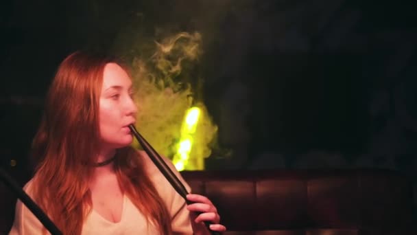 Portré egy nőről, aki vízipipát szív egy társalgóban. A média. Nő hosszú vörös hajjal és fekete fojtogató nyakán pihentető egy shisha bárban. — Stock videók