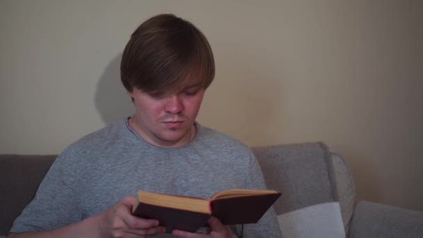 Retrato de um estudante homem concentrado lendo um livro antigo em casa. Conceito. Um homem caucasiano adulto sentado no sofá-cama em casa e estudando o livro. — Vídeo de Stock