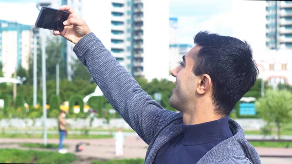 Portret van een vrolijke lachende man die een selfie maakt in het stadspark. De media. Zijaanzicht van een jonge zwarte harige man maken selfie, staande op witte stad gebouwen achtergrond. — Stockfoto