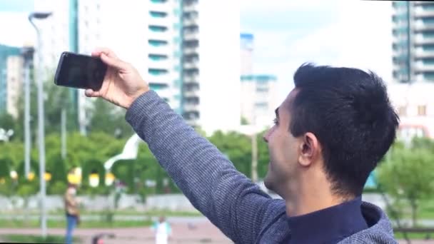 Portret van een vrolijke lachende man die een selfie maakt in het stadspark. De media. Zijaanzicht van een jonge zwarte harige man maken selfie, staande op witte stad gebouwen achtergrond. — Stockvideo