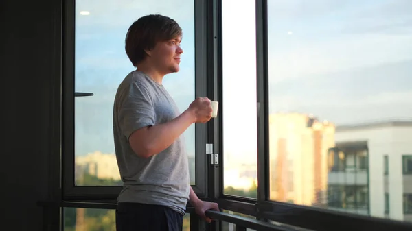 Vue latérale d'un jeune homme heureux tenant une tasse de café sur le balcon à la maison le matin. Concept. Homme buvant du thé ou du café et debout contre une fenêtre ouverte. — Photo