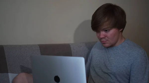 Beau jeune homme assis sur le canapé et utilisant un ordinateur portable dans sa chambre. Concept. Homme blond vêtu d'un t-shirt gris travaillant à la maison, concept d'isolement personnel et freelance. — Photo