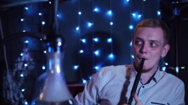 Genç adam mavi çelenk ile süslenmiş karanlık bir odada nargile barında shisha soluyor ve nefes veriyor. Medya. Sarışın adam nargile içiyor ve sağa bakıyor..