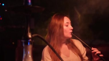 Güzel bir kız kanepede oturuyor ve Shisha tüttürüyor. Medya. Uzun saçlı kadın ağzından ve burnundan beyaz nargile dumanı üflüyor..