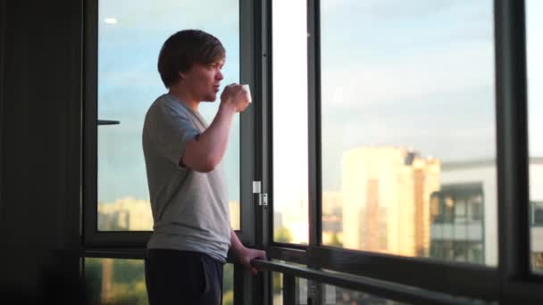 Vista lateral de un joven feliz sosteniendo la taza de café en el balcón en casa por la mañana. Concepto. Hombre bebiendo té o café y de pie contra una ventana abierta. — Vídeo de stock