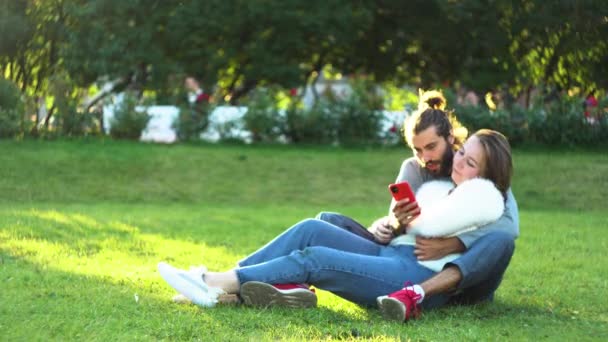 Attractive young couple at the city park sitting on the grass, hugging and looking at smartphone. Concept. Loving man and woman on a romantic date in the summertime. — Stock Video