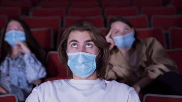 O homem está tensamente assistindo filme no cinema. Mídia. Jovem assiste filme com medo enquanto usa máscara médica. Ir ao cinema durante a pandemia de coronavírus — Vídeo de Stock