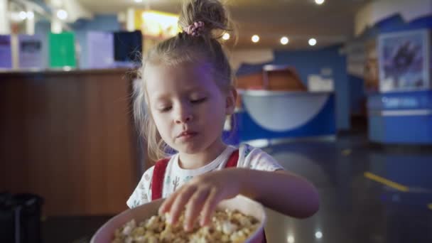 Niña comiendo palomitas. Medios. Linda chica masticando palomitas dulces en el cine. Chica con palomitas de maíz está esperando el comienzo de la caricatura. Ir al cine con niños — Vídeos de Stock