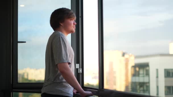 Hombre feliz en casa balcón mirando lejos en la distancia a través de una ventana abierta. Concepto. Vista lateral de un joven rubio disfrutando de la vista urbana. — Vídeo de stock