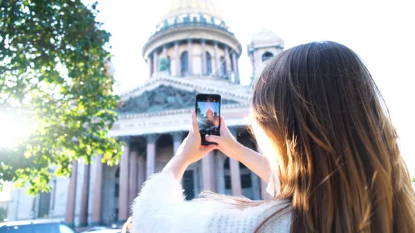 Młoda kobieta na wakacjach fotografuje ze swoim smartfonem piękny zabytkowy budynek. Koncepcja. Widok z tyłu młodego turysty stojącego i korzystającego ze smartfona do robienia zdjęć Świętego Izaaka — Zdjęcie stockowe