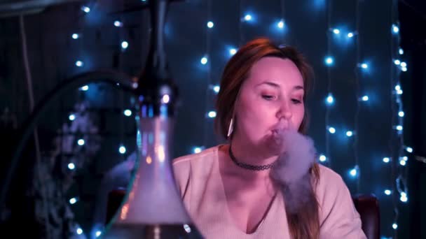 Joven chica hermosa fumando una cachimba en el restaurante. Medios. Retrato de una mujer fumando narguile en el patio de luces azules brillantes, concepto de malos habbits. — Vídeos de Stock