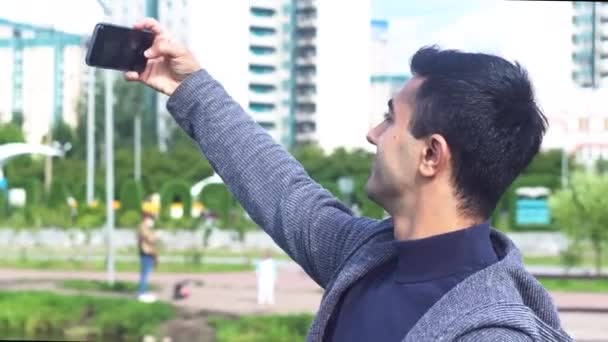 Portret van een vrolijke lachende man die een selfie maakt in het stadspark. De media. Zijaanzicht van een jonge zwarte harige man maken selfie, staande op witte stad gebouwen achtergrond. — Stockvideo