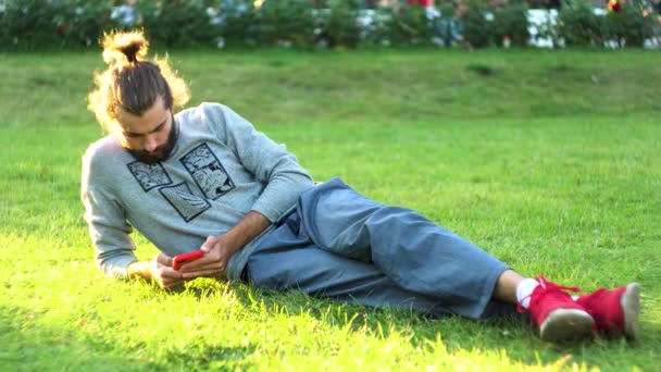 Νεαρός ξαπλώνει στο γρασίδι, ψάχνει σε ένα smartphone, ελέγχει τα κοινωνικά δίκτυα. Έννοια. Λευκός άντρας χαλαρώνει στο πάρκο μόνος του με το κινητό του.. — Αρχείο Βίντεο