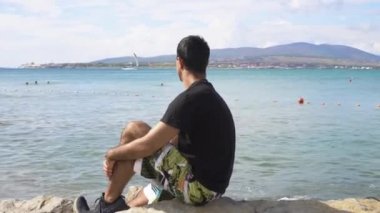 Genç adam taş plajda oturur ve deniz manzarasına bakar. Medya. İnsan denizlerde yüzen insanların arka planında kayanın üzerinde oturur ve ufuktaki dağlarda. Deniz kenarında güneşli bir gün