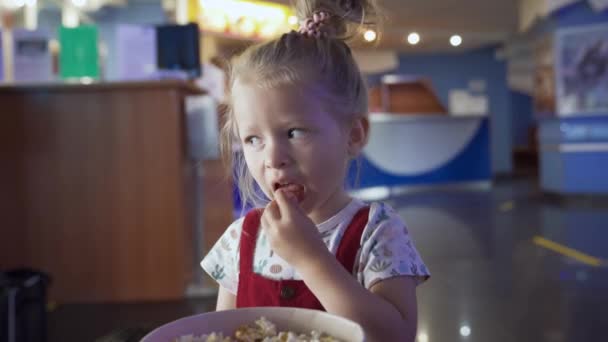 Маленькая девочка ест попкорн. СМИ. Красивая девушка ест попкорн в приемной в кинотеатре. Ребенок ест сладкий попкорн в ожидании мультфильма в кино — стоковое видео