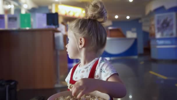 La niña come palomitas de maíz. Medios. Hermosa chica comiendo palomitas en la sala de espera en el cine. Niño come palomitas de maíz dulce mientras espera dibujos animados en el cine — Vídeos de Stock