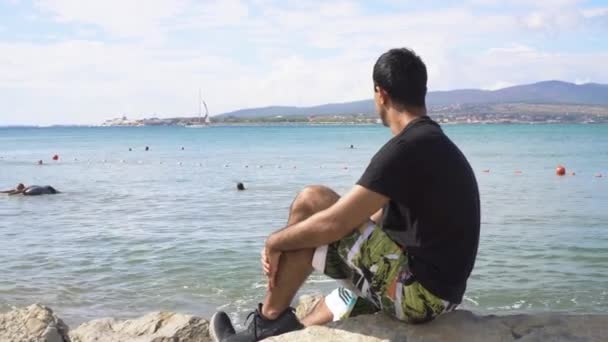 Mladý muž sedí na kamenné pláži a dívá se na moře. Média. Člověk sedí na skále na pozadí lidí vznášejících se v moři a horách na obzoru. Slunečný den po moři — Stock video