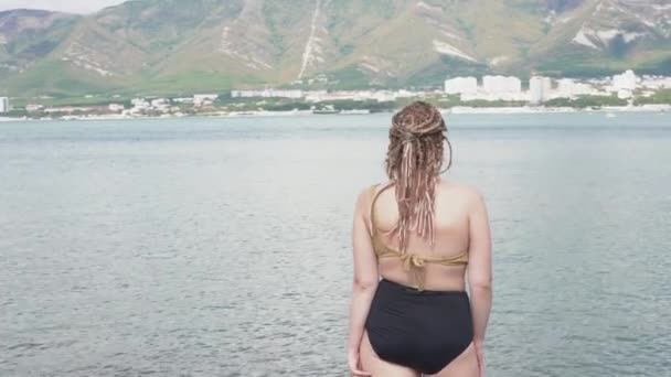 Femme avec des tresses sur fond de mer et de montagnes. Les médias. Vue arrière de la belle femme en maillot de bain et tresses indiennes — Video