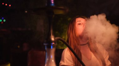 Güzel bir kız kanepede oturuyor ve Shisha tüttürüyor. Medya. Uzun saçlı kadın ağzından ve burnundan beyaz nargile dumanı üflüyor..