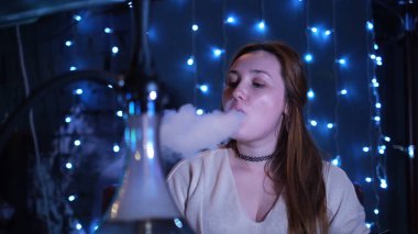 Güzel genç bir kadın burnundan ve ağzından nargile soluyor ve nefes veriyor. Medya. Kızıl saçlı kız mavi çelenkle süslenmiş duvarları olan bir kafede shisha içiyor..