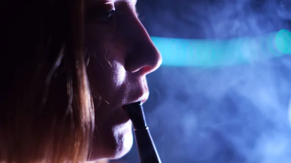 Nő füstölgő shisha egy sötét szobában egy nightclub. A média. Közelkép oldalnézetben egy nő érzéki arc megérinti vízipipipa cső az ajkaival és kilégzés fehér füst. — Stock Fotó