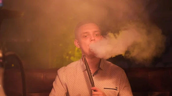 Mann raucht Wasserpfeife vor dunklem Hintergrund einer Nachtbar oder eines Restaurants. Medien. Das Vergnügen, auf dem Gesicht eines jungen Mannes zu rauchen, während er Shisha raucht. — Stockfoto