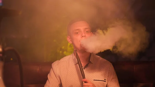 Man rookt een hookah op de donkere achtergrond van een nachtbar of restaurant. De media. Het plezier van roken op het gezicht van een jonge man die tijd doorbrengt terwijl hij shisha rookt. — Stockfoto