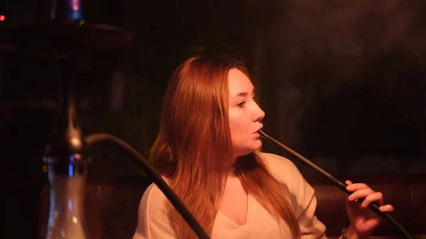 Mooi meisje dat op de bank zit en shisha rookt. De media. Lang haar vrouw uitademen witte rook van hookah door haar mond en neus. — Stockfoto