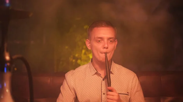 Man rookt een hookah op de donkere achtergrond van een nachtbar of restaurant. De media. Het plezier van roken op het gezicht van een jonge man die tijd doorbrengt terwijl hij shisha rookt. — Stockfoto