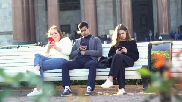 Conceito de vício em smartphones. Mídia. Grupo de amigos, um homem e duas mulheres sentados ao ar livre em um banco no parque e assistindo algo em telefones celulares. — Vídeo de Stock