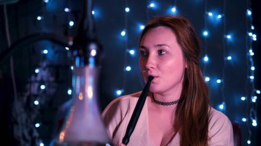 Restoranda ya da gece barında oturup nargile içen güzel bir genç kadın. Medya. Uzun saçlı kadın beyaz duman soluyor ve soluyor, şık doğulu shisha konsepti..