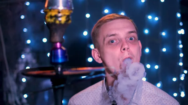 Jonge blonde man met blauwe ogen die rookringen blaast. De media. Man roken hookah aan de nachtbar, het uitademen van rook op blauw schijnende lichten achtergrond. — Stockfoto