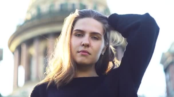 Menina bonita usando óculos de sol sorrindo na rua da cidade no fundo da catedral. Mídia. Retrato de mulher jovem sexy em camisola de malha preta colocando em seus óculos e sorrindo em pé — Vídeo de Stock