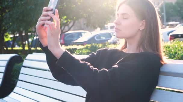 Retrato de uma jovem sob os raios brilhantes do sol de verão usando telefone inteligente. Mídia. Mulher bonita fazendo selfie enquanto sentado em um banco branco em um parque da cidade. — Vídeo de Stock