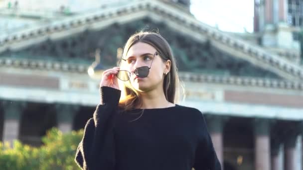 Mooi meisje met een zonnebril lachend in de stad straat op de achtergrond van de kathedraal. De media. Portret van sexy jonge vrouw in zwart gebreide trui zetten op haar bril en glimlachen terwijl staan — Stockvideo