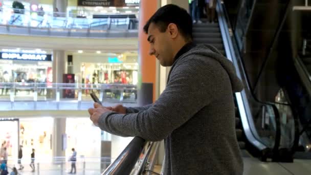 Seitenansicht eines jungen Mannes, der im Einkaufszentrum steht und sein Smartphone benutzt. Medien. Mann in Freizeitkleidung textet vor dem Hintergrund einer fahrenden Rolltreppe. — Stockvideo