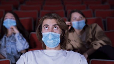Adam yoğun bir şekilde sinemada film izliyor. Medya. Genç adam korku içinde tıbbi maske takıp film izliyor. Koronavirüs salgını sırasında sinemaya gitmek