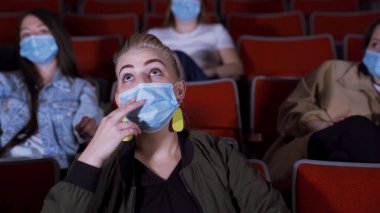 Kadın koruyucu maskeyle film izliyor. Medya. Güzel, genç bir kadın, sinemayla ilgilenen bir film izliyor. Koronavirüs salgını sırasında sinemaya gitmek