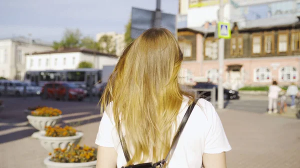 Vista posterior de mujer joven con el pelo revoloteando en el viento. Medios. Hermosa joven con el pelo dorado pasea por la ciudad en tiempo soleado. Paseando por la ciudad — Foto de Stock