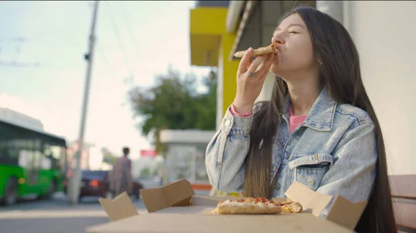 Piękna kobieta je pizzę na ulicy. Media. Kobieta podjadająca pizzę przed kawiarnią. Młoda kobieta je pizzę na tle ruchliwego miasta — Zdjęcie stockowe