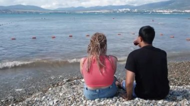 Genç bir çift sahilde şarap içiyor. Kavram. Kız arkadaşı denizde rahatlayan erkek arkadaş görüntüsü. Kumsalda oturmuş şarap içen iki güzel sevgili