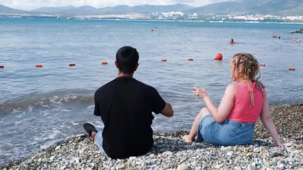 Mladý pár pije víno na pláži. Koncept. Zadní pohled na přítele s přítelkyní relaxující na moři. Krásný pár milenců sedících na pláži a pijících víno — Stock video
