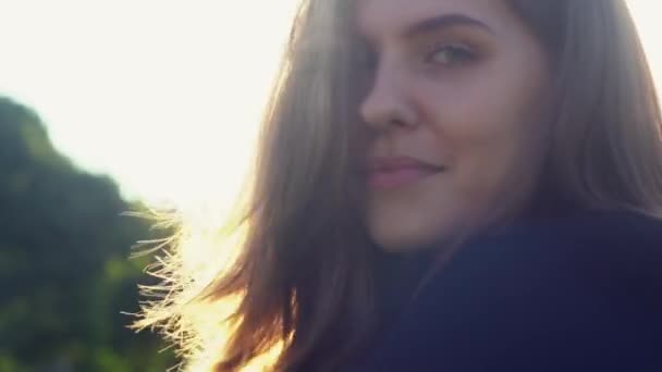 Impresionante cara de chica contra el sol con su pelo soplando en el viento. Medios. Primer plano de una joven posando al aire libre, sonriendo a la cámara y tocándose el pelo. — Vídeos de Stock