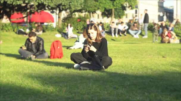 Vrouw in zwarte kleren zittend op groene weide van een stadspark. De media. Concept van slechte gewoonte, jong meisje roken elektronische sigaret en kijken naar haar smartphone. — Stockvideo