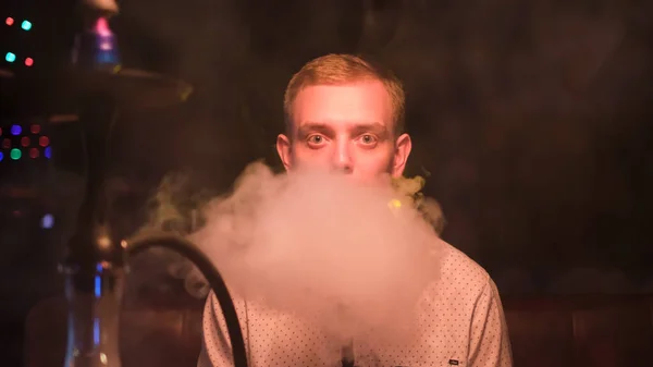 Jonge blonde man met grote groene ogen die shisha rookt in restaurant. De media. Man rustend met hookah recht naar de camera kijkend laat in de avond. — Stockfoto